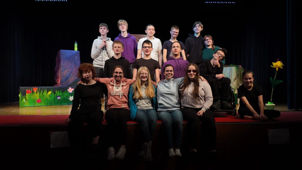 Gruppenfoto der Schauspielenden, in mehreren Reihen auf der Bühne sitzend bzw. stehend. Dahinter Teile des Bühnenbilds.