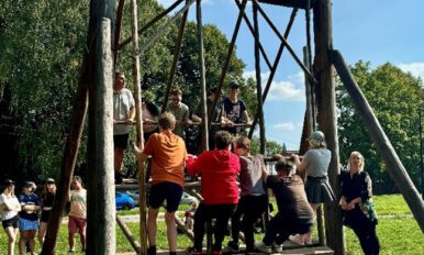 **Alternativtext:** Eine große Holzschaukel auf einer Wiese, die von mehreren Personen gemeinsam genutzt wird. Einige stehen auf der Plattform der Schaukel, während andere am Boden stehen und schaukeln. Im Hintergrund sind Zuschauer, Bäume und ein blauer Himmel zu sehen. Die Szene vermittelt ein Gefühl von Gemeinschaft und Aktivität an der frischen Luft.