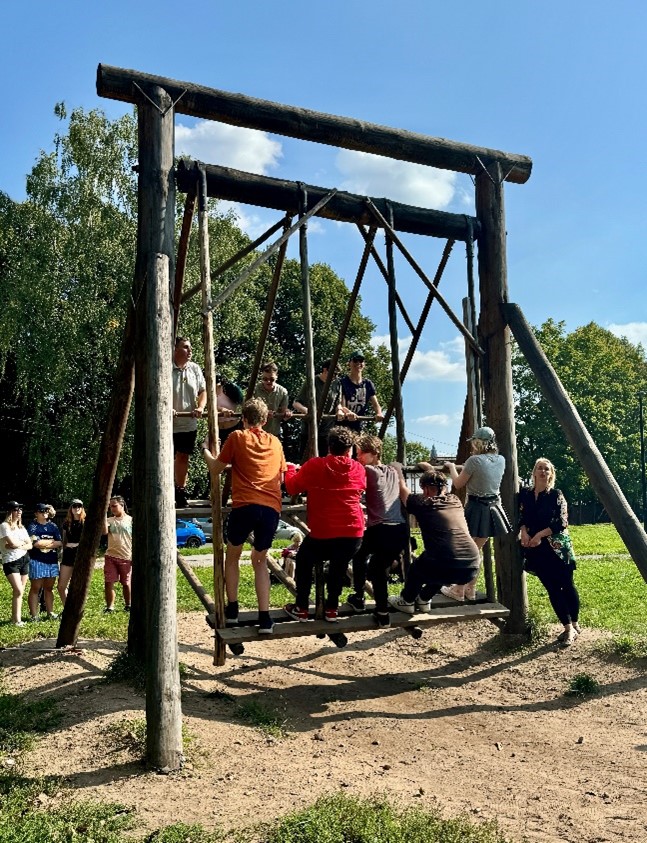 **Alternativtext:** Eine große Holzschaukel auf einer Wiese, die von mehreren Personen gemeinsam genutzt wird. Einige stehen auf der Plattform der Schaukel, während andere am Boden stehen und schaukeln. Im Hintergrund sind Zuschauer, Bäume und ein blauer Himmel zu sehen. Die Szene vermittelt ein Gefühl von Gemeinschaft und Aktivität an der frischen Luft.